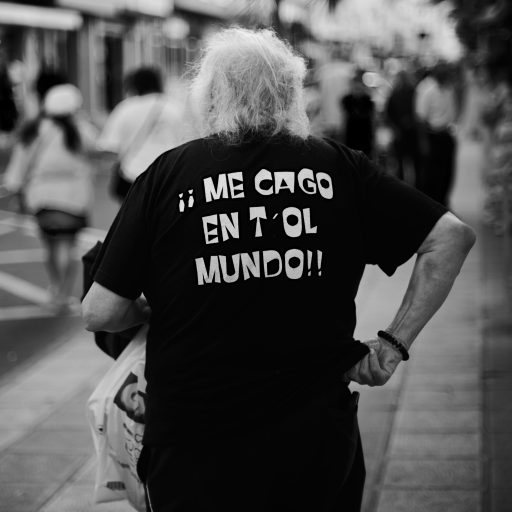 Hombre de pelo blanco de espaldas con una camiseta que dice “¡¡Me cago en t’ol mundo!!”.