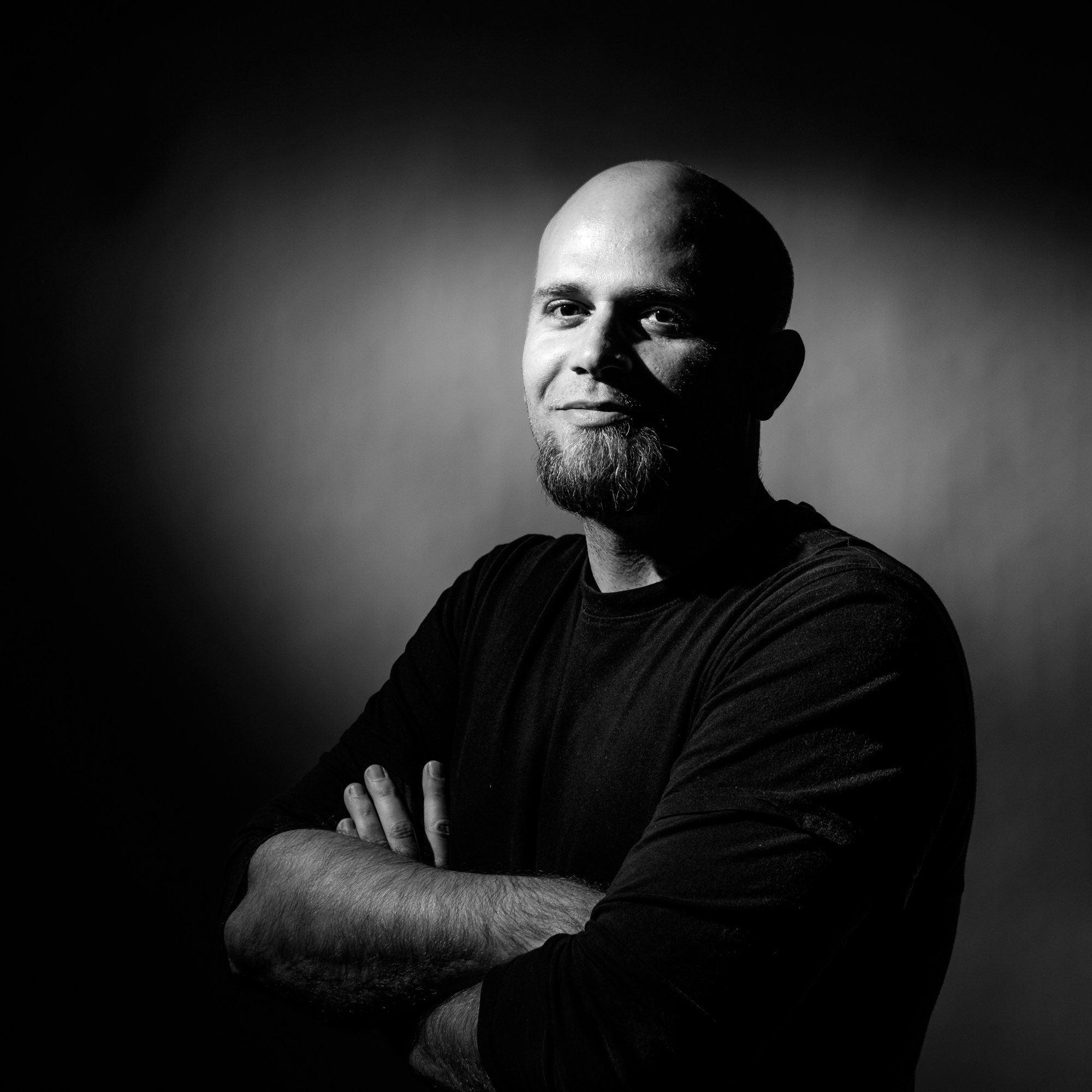 Retrato de hombre calvo con barba y los brazos cruzados, iluminado sobre fondo oscuro.