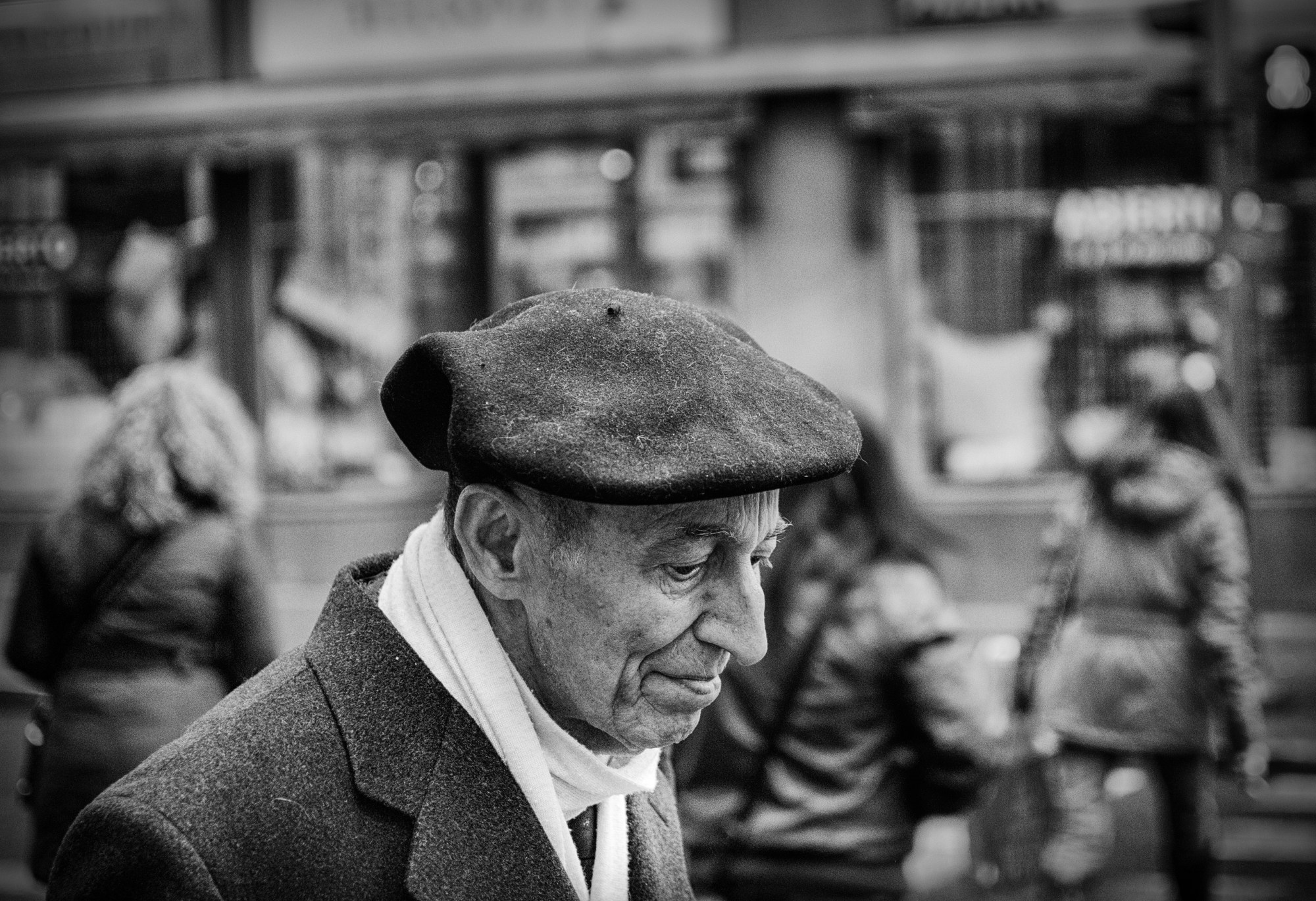 Hombre mayor con boina y bufanda blanca, de perfil, en una calle concurrida.