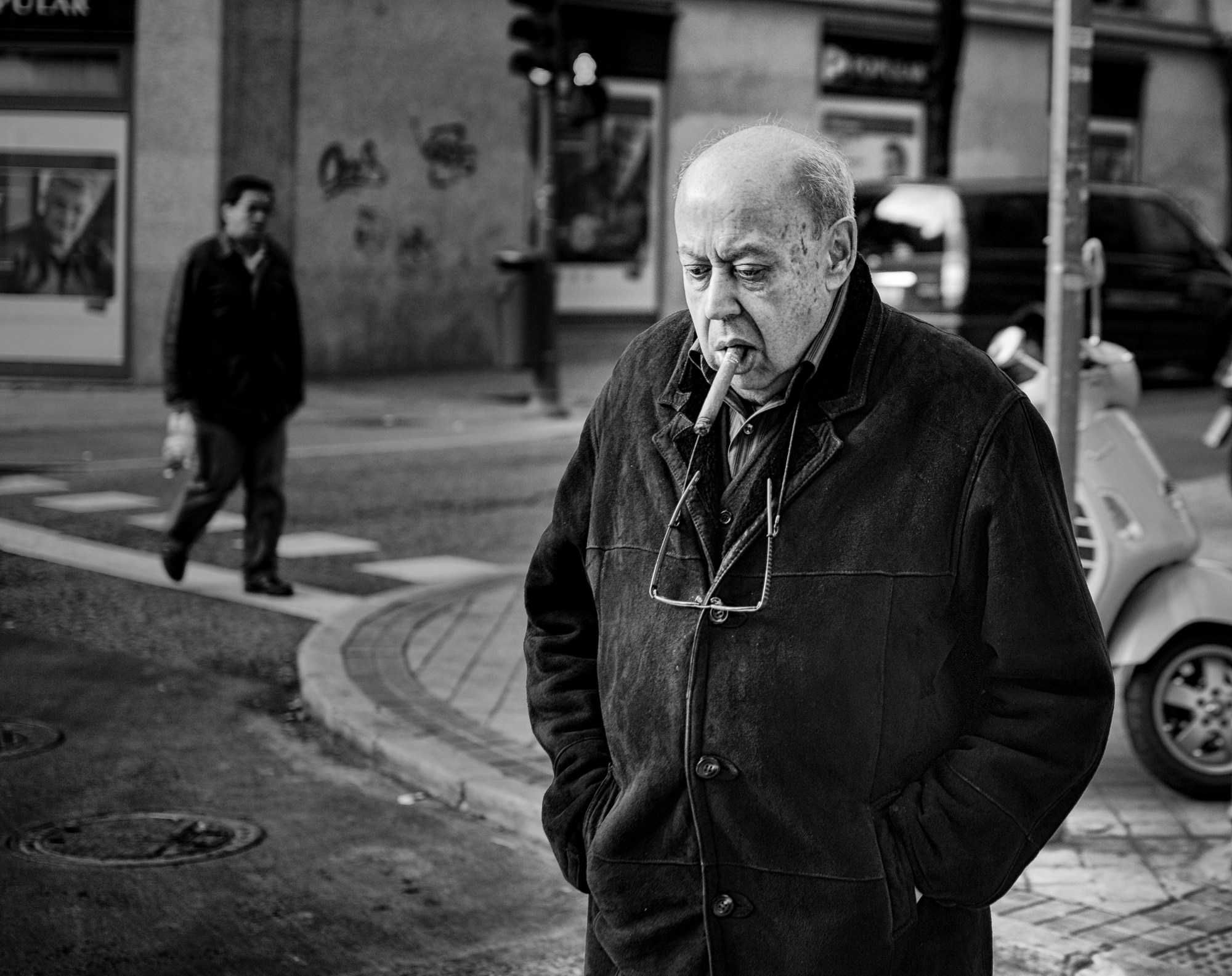Hombre calvo con abrigo y un puro en la boca, con las manos en los bolsillos, en una esquina.