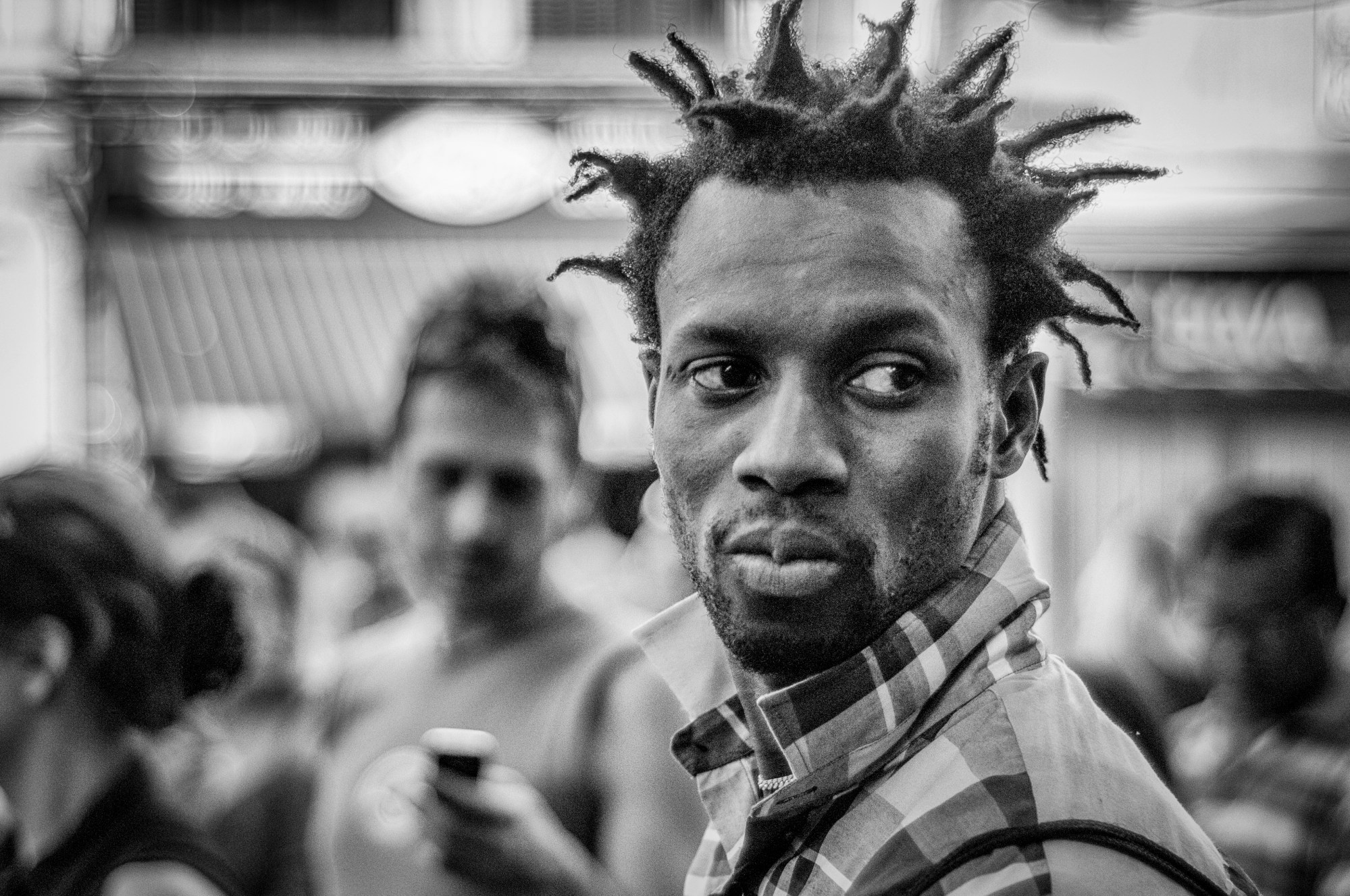Hombre con rastas cortas y camisa de cuadros mirando de reojo, con gente desenfocada detrás.