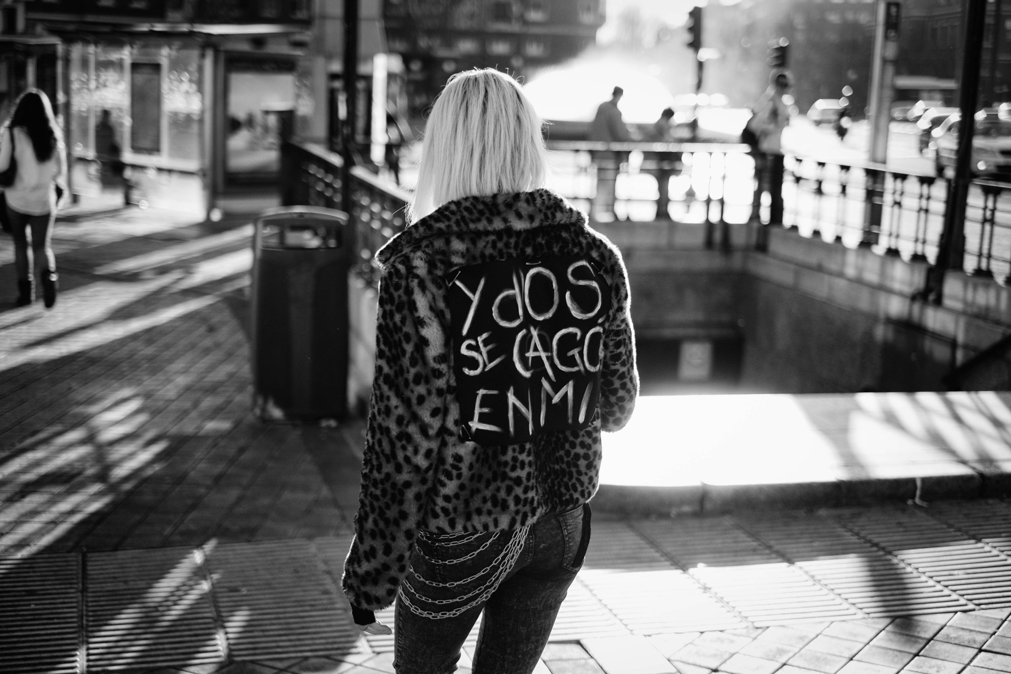 Mujer rubia de espaldas con chaqueta de leopardo y un texto pintado a mano en la espalda, junto a una boca de metro.