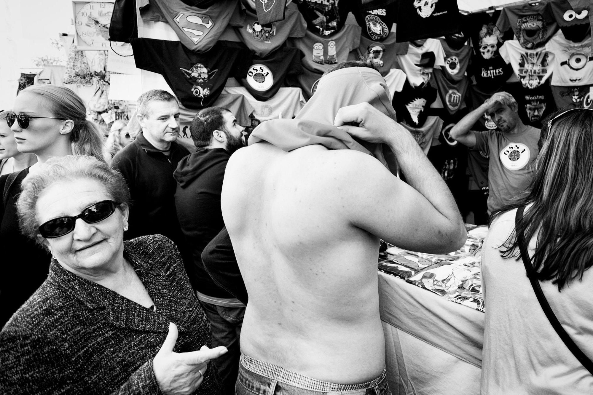 Hombre sin camisa quitándose una prenda ante un puesto de camisetas, entre una mujer con gafas de sol y otros transeúntes.