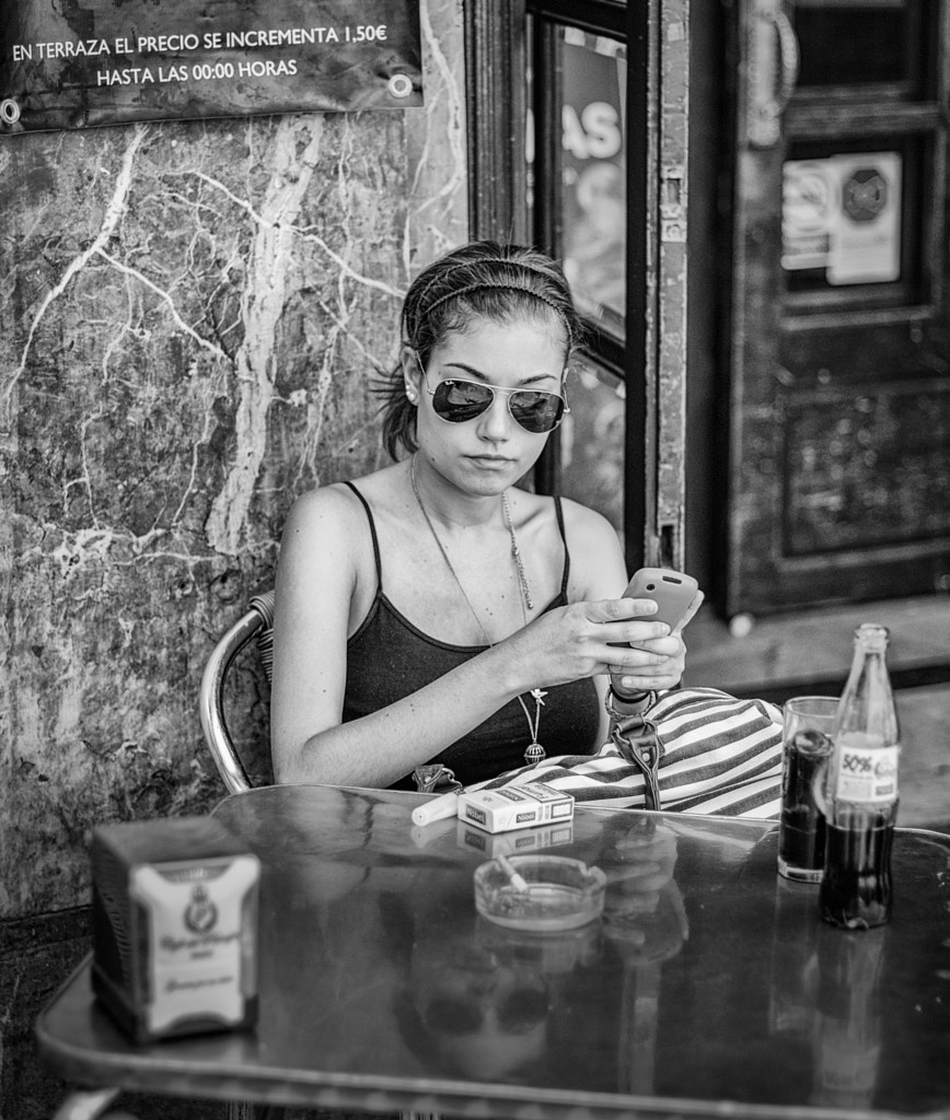 Mujer joven con gafas de sol mirando el móvil en la mesa de una terraza, con una botella de refresco.