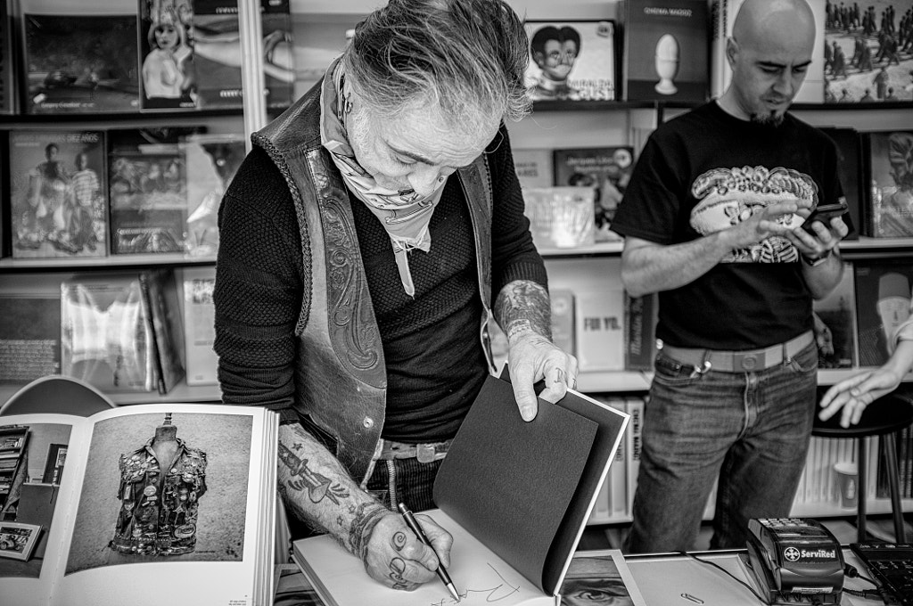 Hombre tatuado con pañuelo al cuello firmando un libro en una librería, junto a otro hombre que mira el móvil.