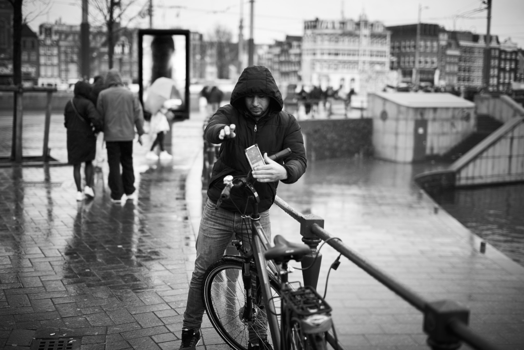 Joven con capucha junto a una bicicleta en una calle mojada, con edificios al otro lado de un canal.