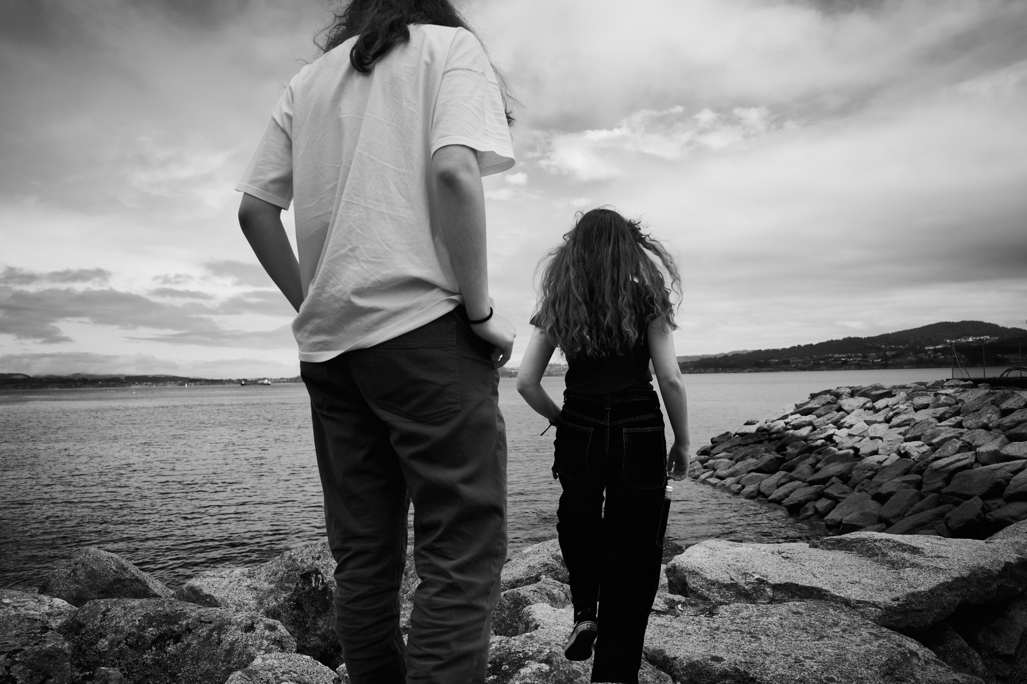 Dos personas de espaldas sobre unas rocas mirando al mar bajo un cielo nublado.