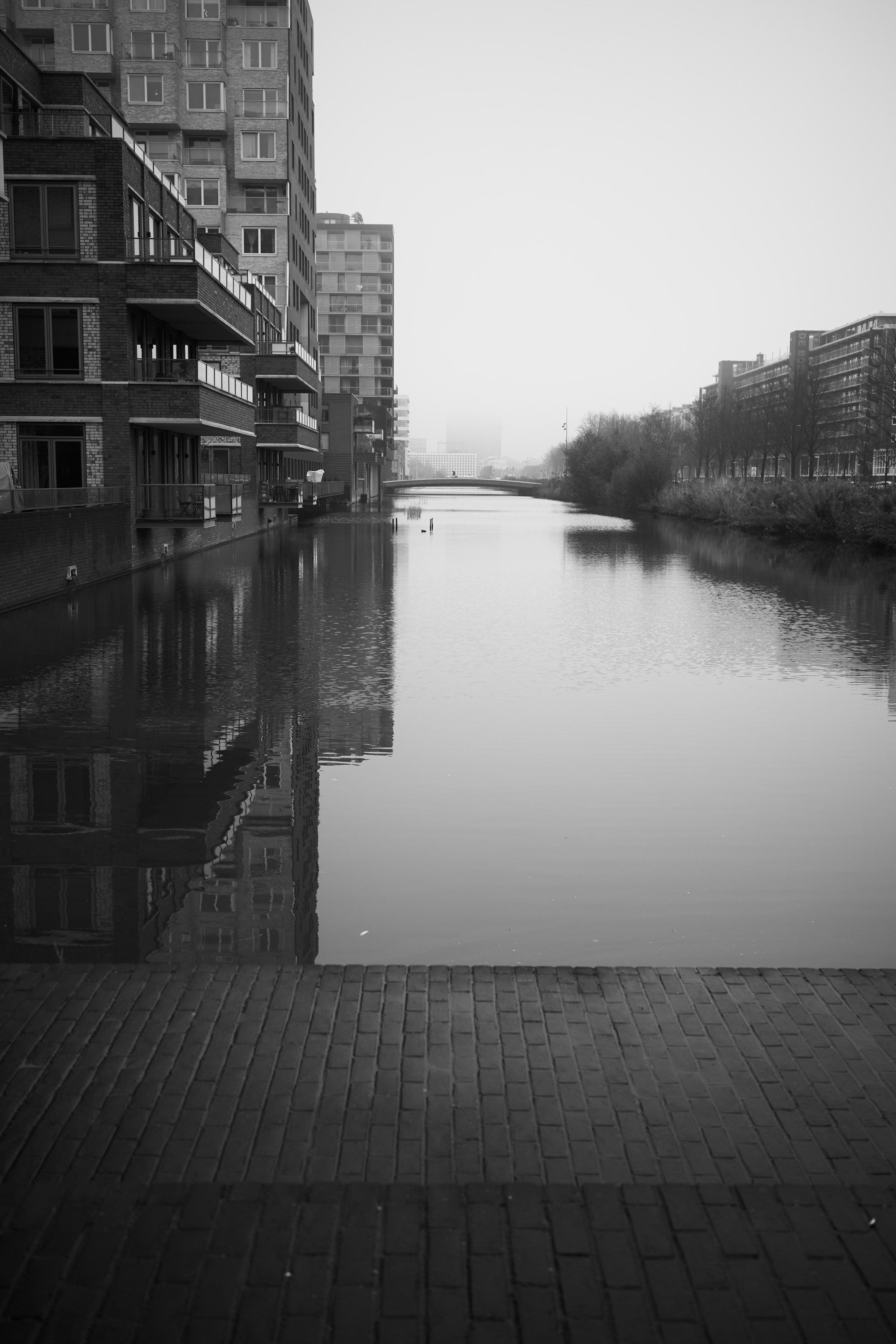 Canal en calma entre edificios de viviendas, con niebla al fondo.