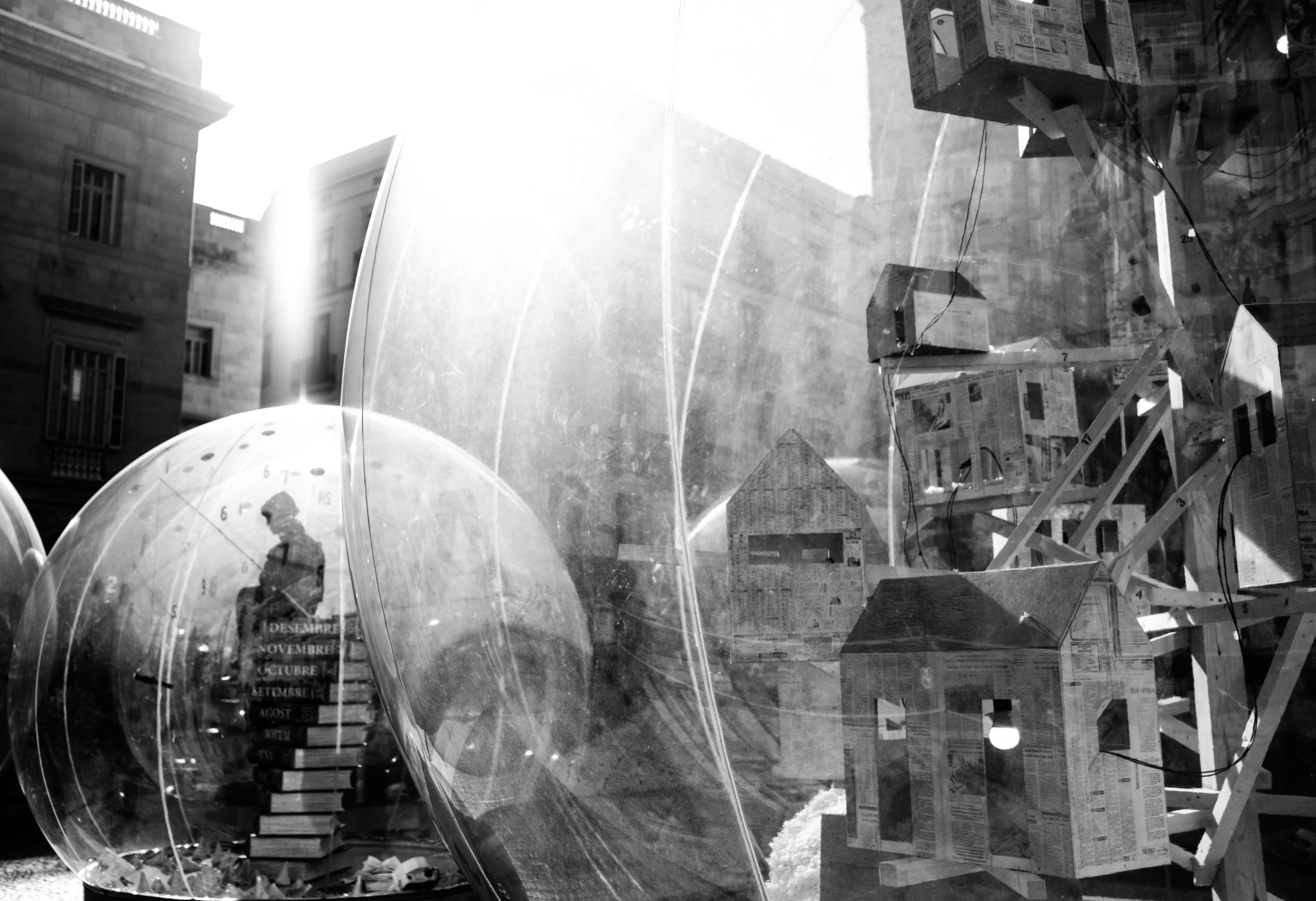 Esferas transparentes y una estructura de casitas apiladas en una plaza, a contraluz.