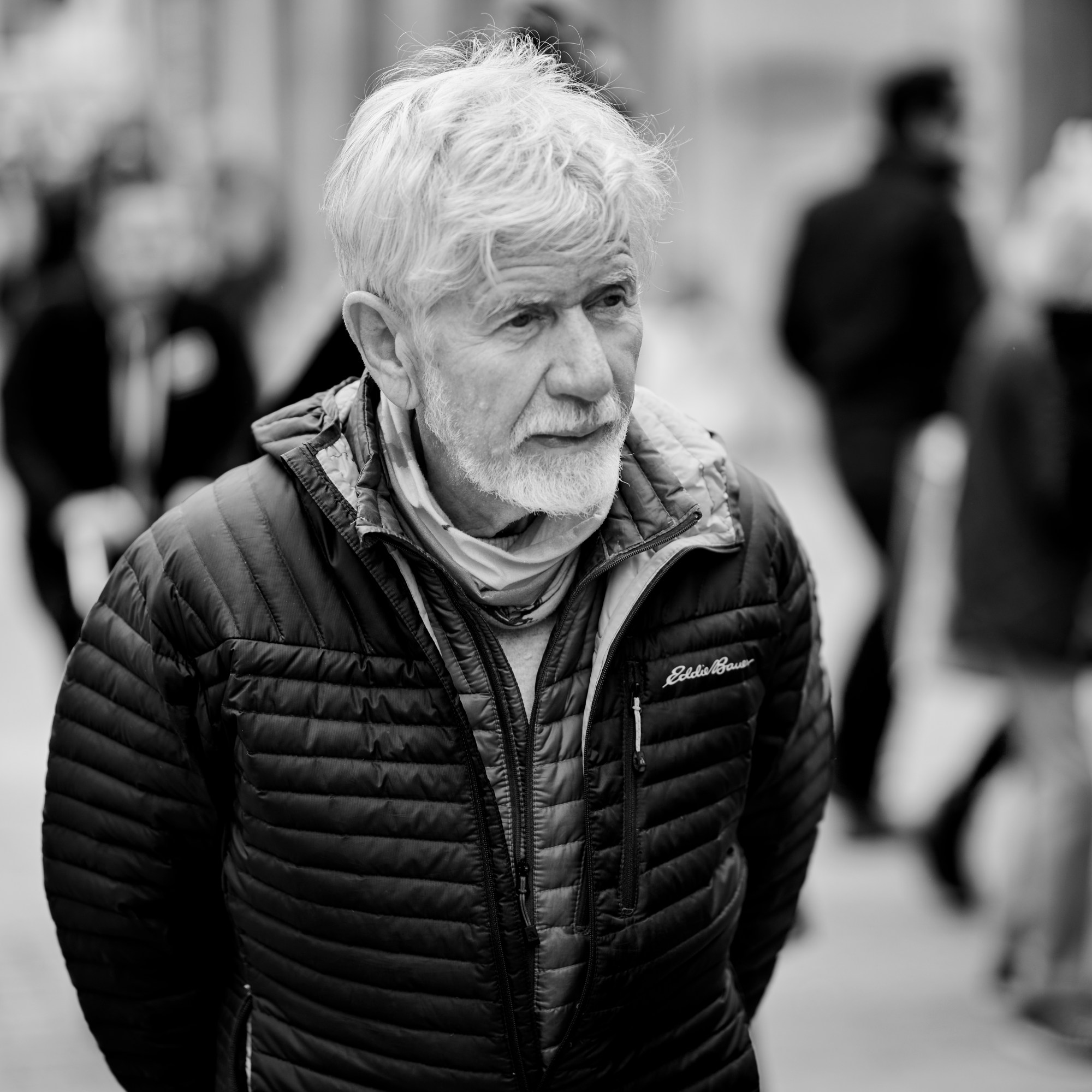 Hombre de pelo y barba blancos con plumífero, entre peatones desenfocados.