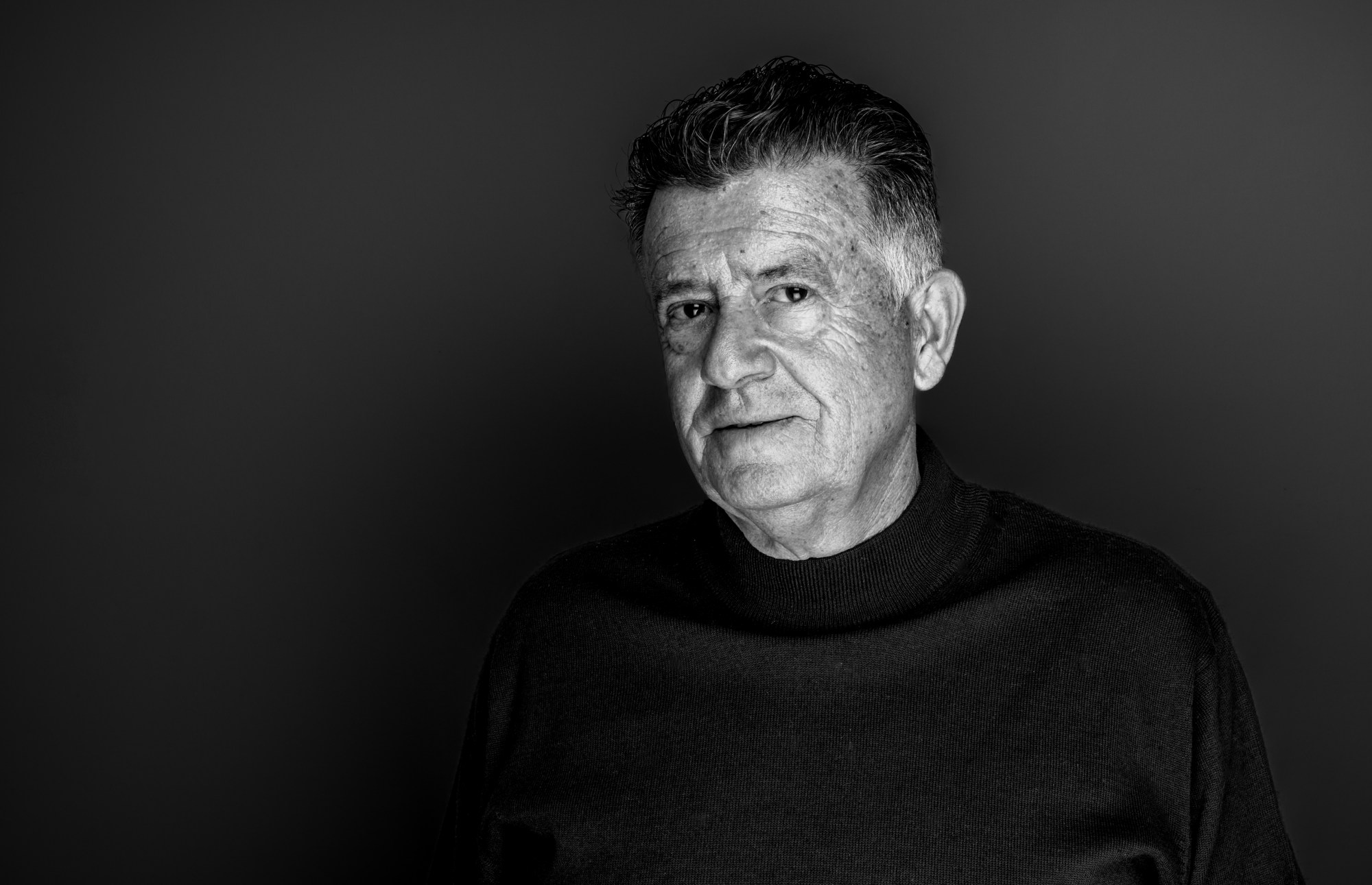 Retrato de hombre mayor con jersey oscuro sobre fondo oscuro.