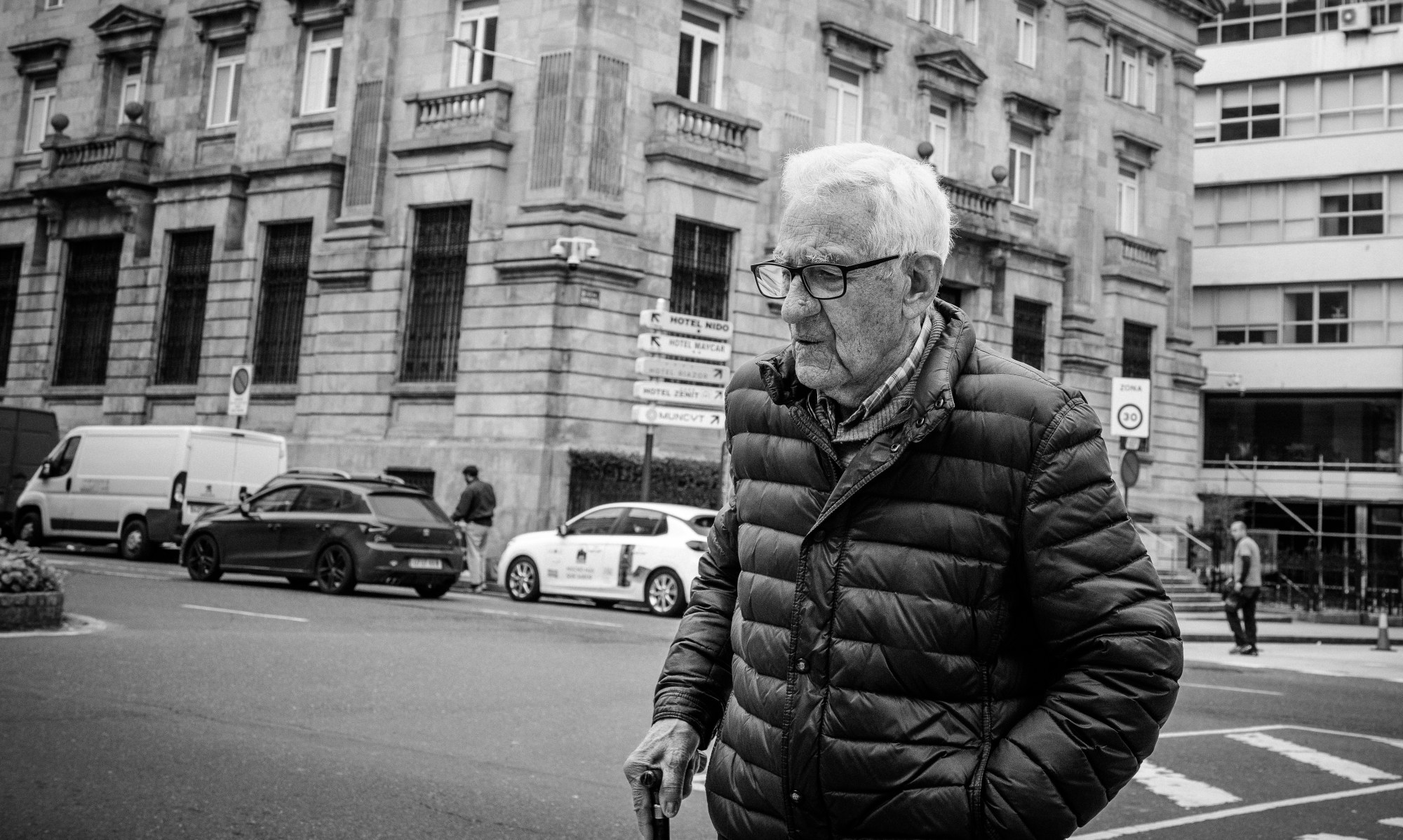 Hombre mayor con gafas y plumífero cruzando una calle con edificios de piedra al fondo.