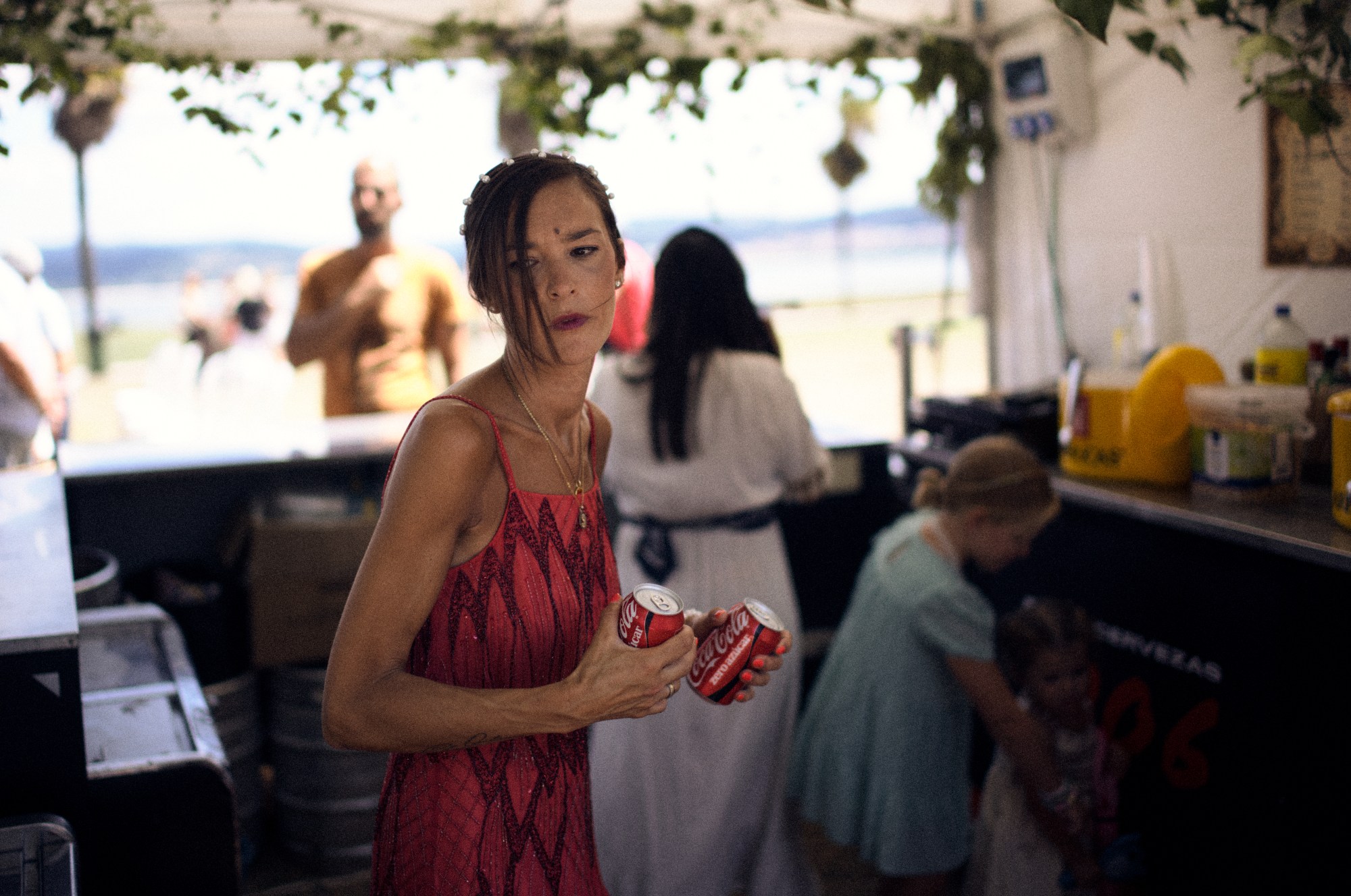 Mujer con vestido rojo sosteniendo dos latas de refresco en un puesto de bebidas, con el mar al fondo.