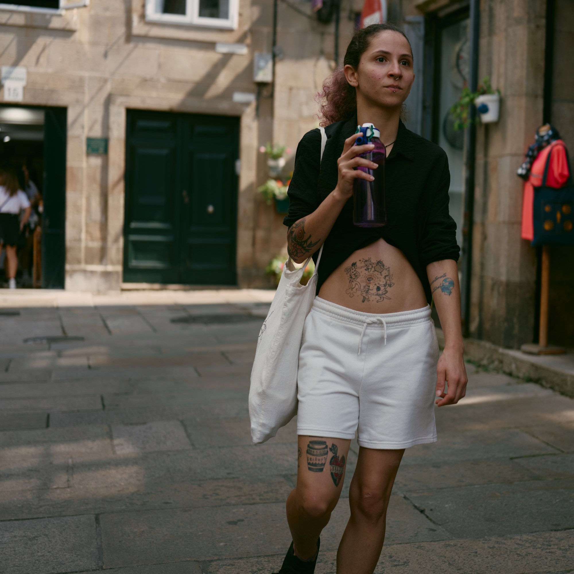 Mujer joven con camisa negra y pantalón corto blanco caminando con una lata en la mano por una calle de piedra.
